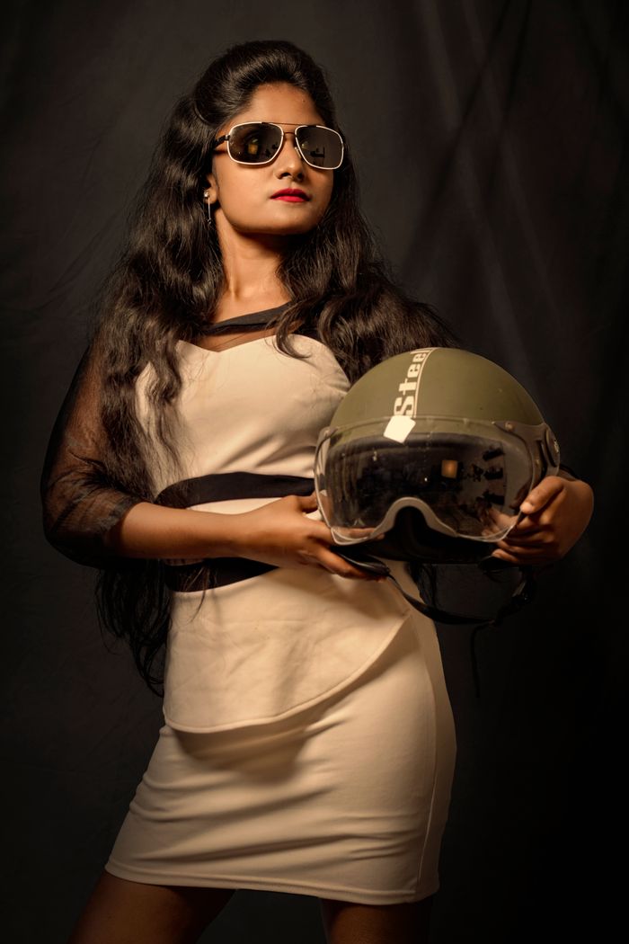Glamorous young woman in dress and sunglasses posing with a helmet in a studio setting.